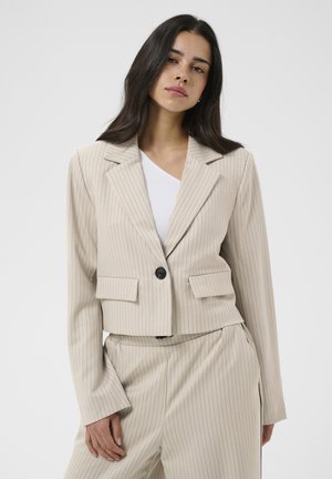 Pikkade tumedate juustega naine, kes kannab beežika pinstripe lühikest jopet ja sellele sobivaid kõrge vöökohaga pükse ühtlasel heledal taustal.