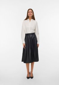 Chemise blanche à boutons, fabriquée en tissu léger, associée à une jupe midi en simili cuir noir plissée. La tenue est complétée par des talons noirs.