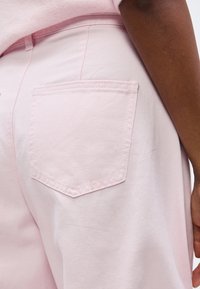 Κοντινό πλάνο ενός ατόμου που φοράει ανοιχτό ροζ παντελόνι με πίσω τσέπη, εμφανίζοντας μέρος του χεριού και του κορμού του σε λευκό φόντο.