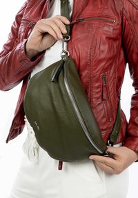 Borsa a tracolla verde in pelle con forma curva, cerniera argentata e tracolla regolabile, indossata sopra una giacca rossa in pelle. Texture liscia con branding minimale.