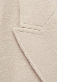 Tessuto di lana beige con una texture morbida, caratterizzato da un dettaglio di cucitura angolare. Il materiale appare spesso e caldo, adatto per capi d'abbigliamento esterni.