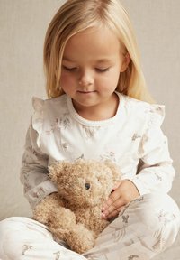 Barn i lyse blomstermønstrede pyjamas med flæser, holder en beige, fluffy bamse mod en neutral baggrund.