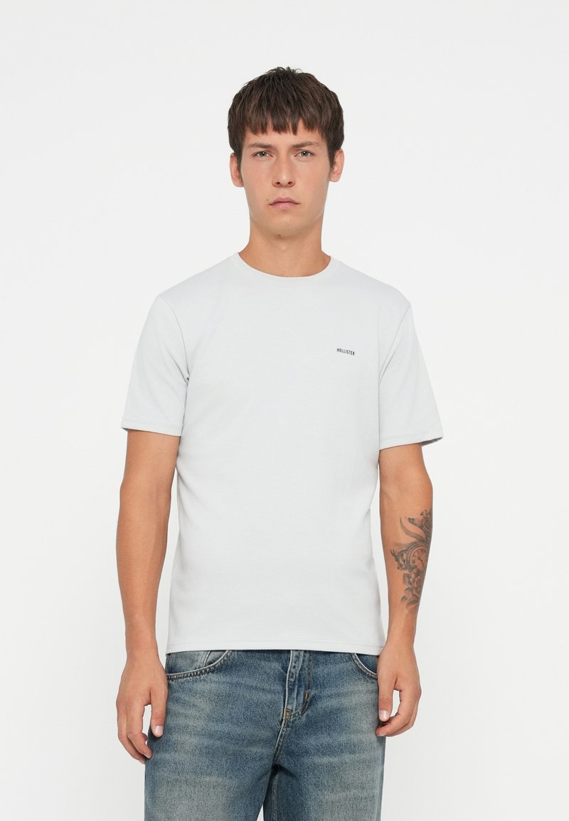 Camiseta gris claro con escote redondo y mangas cortas, que presenta un pequeño logo de "Hollister" en el lado izquierdo del pecho. Textura de tela suave.