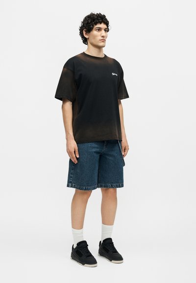 Νέος άνδρας με σγουρά μαλλιά που φοράει μαύρο ξεθωριασμένο μπλουζάκι, μπλε τζιν σορτς, λευκές κάλτσες και μαύρα αθλητικά παπούτσια, στέκεται μπροστά σε απλό φόντο.