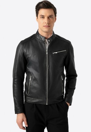 Chaqueta de cuero negra con cuello alzado, cierre frontal de cremallera y bolsillos con cremallera. Textura suave con diseño ajustado y acentos en hardware plateado.