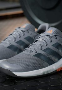 Zapatillas deportivas grises con una parte superior de malla texturizada, detalles negros y toques naranjas. Presentan cordones visibles y una suela acolchada.