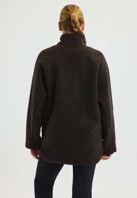 Nainen, jolla on letitetyt hiukset, pukeutuneena tummanruskeaan fleecetakkiin ja mustiin housuihin, kääntynyt pois pelkää valoisaa taustaa vasten.