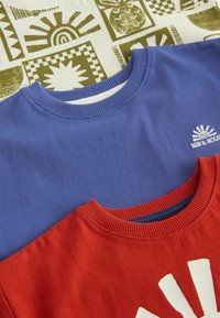 Deux sweat-shirts : un bleu avec un col côtelé et un petit logo blanc ; un rouge avec un col côtelé et un motif d'éclat de soleil blanc.
