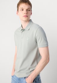 Lys grå polo med krage, korte ermer og en liten logo på brystet. Glatt stoff, avslappet passform, kombinert med lyseblå jeans.