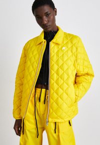 Casaco amarelo acolchoado com colarinho, apresentando um fecho de correr branco e logótipo. Combinado com calças amarelas a condizer e uma camada interior preta. Textura suave.