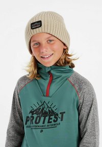 Cappello a coste beige con un patch del logo, abbinato a un top verde con zip decorato da una grafica montana e maniche grigie.