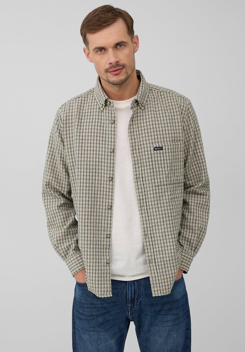 Man in een beige geruite button-up shirt over een wit T-shirt, blauwe jeans, staande met zijn handen in de zakken tegen een effen achtergrond.