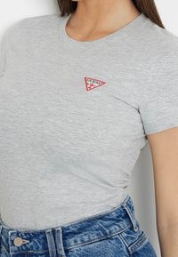 Femme portant un t-shirt gris clair à manches courtes avec un petit logo Guess rouge et blanc sur la poitrine et un jean bleu.