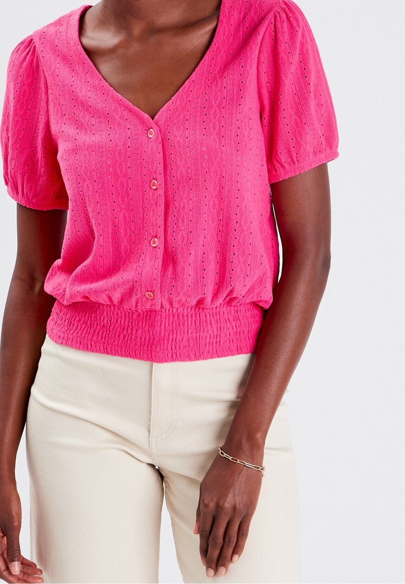 Femme portant un chemisier à manches courtes boutonné rose vif en tissu texturé et un pantalon beige, debout devant un fond uni.