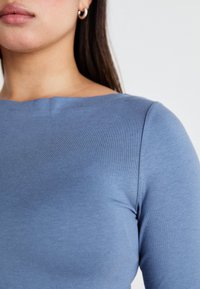 Μπλε εφαρμοστό τοπ φτιαγμένο από έναν λείο, ελαστικό ύφασμα, με στρογγυλή λαιμόκοψη και διακριτές ραφές στους ώμους.