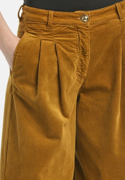 Cord-Hosen in einem satten Senfton, mit vorderen Falten, einem Knopfverschluss und seitlichen Taschen. Die Textur zeigt feine Rippungen.