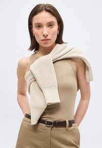 Νεαρή γυναίκα που φοράει ένα αμάνικο μπεζ τοπ και καφέ παντελόνι με ένα κρεμ πλεκτό πουλόβερ που είναι περασμένο πάνω από τους ώμους της, μπροστά σε απλό φόντο.