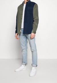 Napillinen paita, jossa on tummansininen ja oliivinvihreä väriblokkimalli, tehty corduroysta ja jossa on rintatasku. Yhdistetty vaaleansinisiin farkkuihin ja valkoisiin tennareihin.