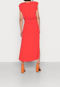 Robe maxi rouge avec manches courtes évasées, taille élastique et fentes latérales. Associée à des sandales à talons blocs orange. Texture de tissu lisse.