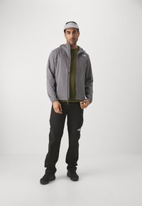 Grijze zip-up jas met capuchon en borstzak, groene shirt eronder, zwarte broek en donkere schoenen. Pet heeft een logo en is lichtgrijs van kleur.