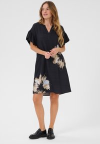 Schwarzes Kleid mit V-Ausschnitt, kurzen Rüschensäumen und einem floralen Muster in Creme und Gold im unteren Bereich. Getragen mit schwarzen Schuhen.