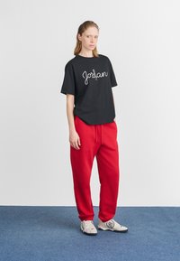 Maglietta nera con la scritta "Jordan" in bianco, abbinata a pantaloni della tuta rossi e scarpe grigie. La maglietta ha una vestibilità ampia e maniche corte.