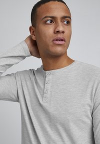 Lys grå langermet henley-skjorte med rund hals, tre knapper foran og teksturert stoff. Glatt overflate, avslappet passform.