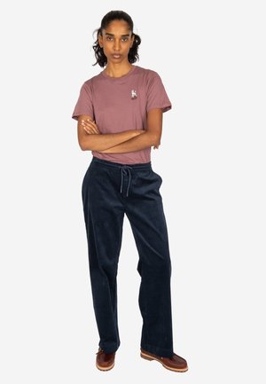 Woman with folded arms wearing a mauve t-shirt, dark blue pants, and brown shoes, standing against a white background.
