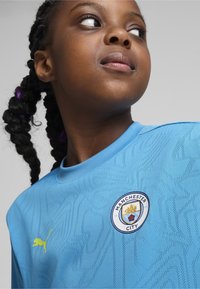 Vaaleansininen jalkapalloserti, jossa on teksturoitu kuvio, keltainen Puma-logo ja Manchester City -symboli rinnassa.