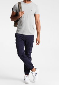 Graues Rundhals-T-Shirt, marineblaue Cargo-Hosen mit Taschen, weiße Sneakers und einen beigefarbenen Rucksack tragend, vor einem schlichten Hintergrund stehend.