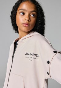 Svijetlo ružičasta zip-up dukserica od pamuka. Ima otisak logotipa, velike prednje džepove i kontrastne crne dugmadi na rukavima.