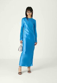 Mulher vestindo um longo vestido azul brilhante, sapatos de salto preto com tiras, uma pequena bolsa prateada e óculos de sol escuros, de pé contra um fundo claro e simples.
