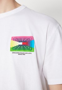 Camiseta de algodón blanca con un gráfico rectangular multicolor que presenta el texto "Tommy Jeans" y el eslogan "Difundiendo paz y amor desde 1985".