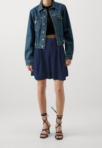 Jeansjacka med en kortad design, mörkblå färg, med knappdetaljer, ihop med en marinblå utsvängd kjol och svarta sandaletter med remmar.