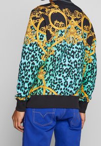 Sudadera negra y turquesa con estampado de leopardo y patrones dorados ornamentales. Incluye detalles de puños y dobladillo acanalados, combinada con pantalones azules.