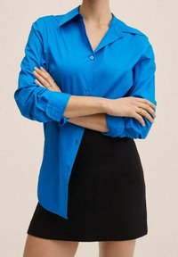 Chemise bleue à boutons en tissu lisse, dotée d'un col et de manches longues retroussées. Portée avec une jupe noire texturée.