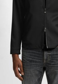 Schwarze Jacke mit Reißverschluss und hohem Kragen, aus glattem Material gefertigt. Kombiniert mit dunklen Jeans, die einen geraden Schnitt zeigen.