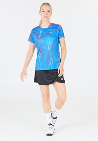 Blå och rosa idrottströja med dynamiska mönster, kombinerad med en svart kjol. Utan logotypsdetalj och vita strumpor med svarta accenter.