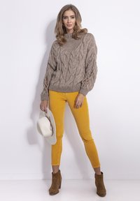 Beżowy sweter w warkoczowy splot z ribbowymi detalami, zestawiony z piaskowymi spodniami z kordurowej tkaniny. Model trzyma kremowy kapelusz. Brązowe botki sięgające kostki dopełniają stylizację.
