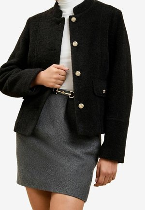 Femme portant une veste noire texturée avec des boutons dorés, un col roulé blanc, une ceinture noire et une jupe en laine grise, montrée du cou jusqu'à mi-cuisse.