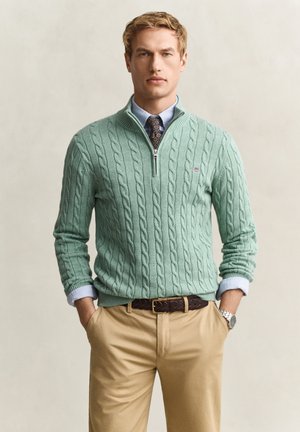 Junger Mann in einem hellgrünen Kabelstrick-Quarter-Zip-Pullover über einem blauen Hemd und gemusterter Krawatte, beigen Hosen und braunem Gürtel, Hände in den Taschen.