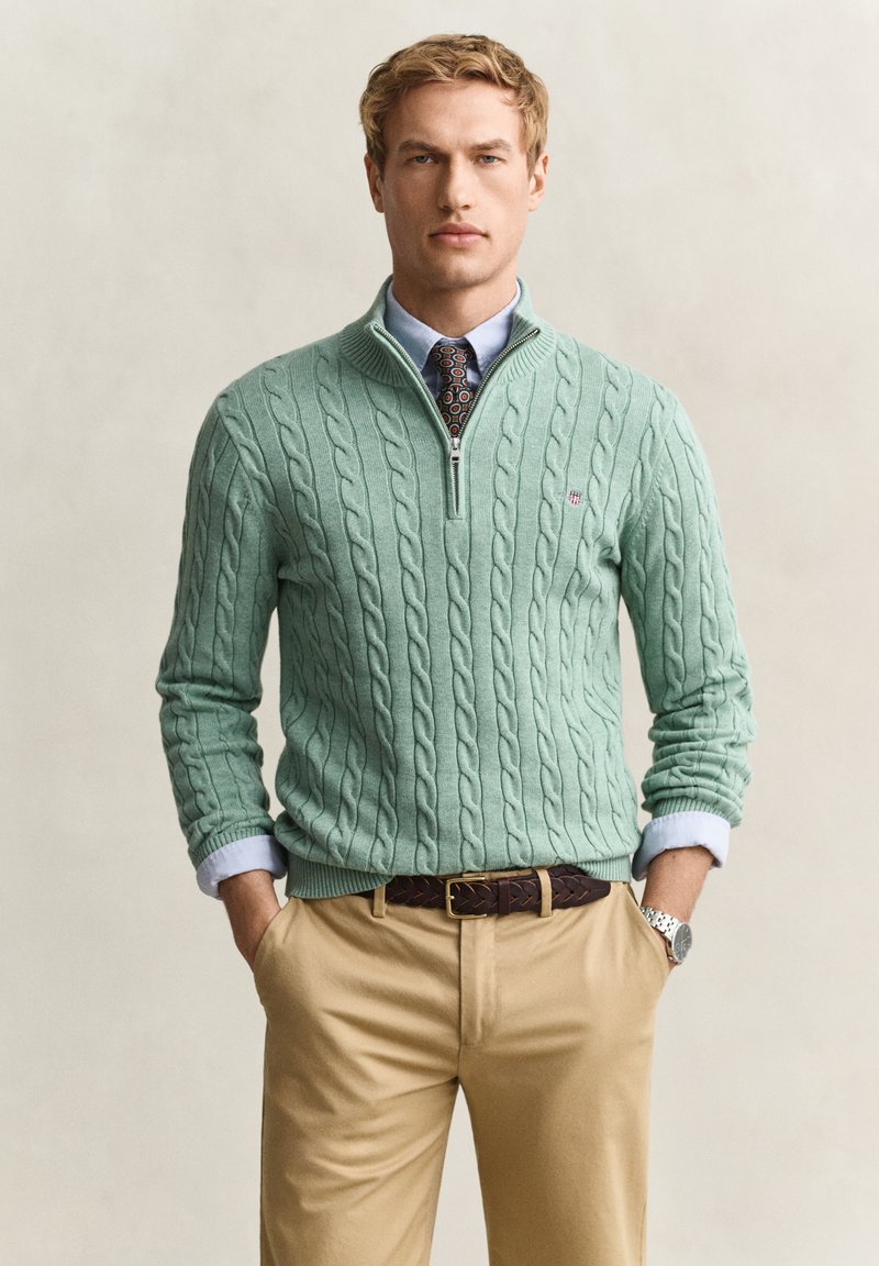 Jeune homme portant un pull en maille torsadée vert clair avec un quart de zip par-dessus une chemise bleue et une cravate à motifs, pantalon beige et ceinture marron, mains dans les poches.