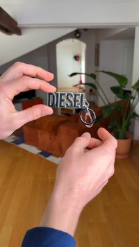 Keychain with "DIESEL" in bold, glossy black letters, attached to a silver metal ring and clip. Background features wooden flooring and a plant.