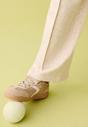Foot wearing beige sneaker and light pants stepping on a yellow tennis ball on a light green surface.