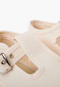 Písková plátěná bota s designem T-strap, kovovou sponou a otevřenými stranami. Obsahuje detaily šití a texturovaný povrch.