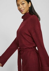 Pull en col roulé bordeaux avec une coupe décontractée, des manches longues et une taille nouée. Fabriqué en tissu doux et texturé avec des manches larges.