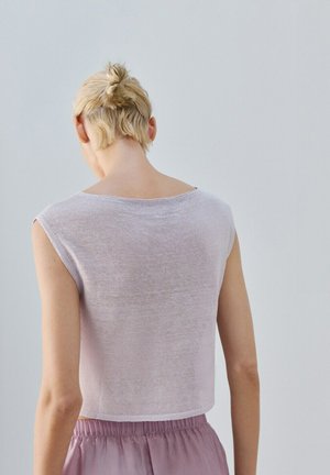 Oseba s kratkimi blond lasmi v figi, oblečena v svetlo vijoličast brezrokavnik in vijolične hlače z elastičnim pasom, obrnjena stran proti enobarvnemu ozadju.