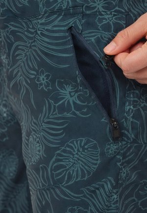 Hand zieht eine kleine Reißverschlusstasche an dunkelblauen Stoffhosen mit floralem Blattmuster auf.