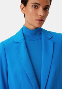 Helderblauwe blazer met gestructureerde revers over een bijpassende coltrui. Gladde stof met minimale textuur, nauwsluitend ontwerp voor een op maat gemaakte uitstraling.