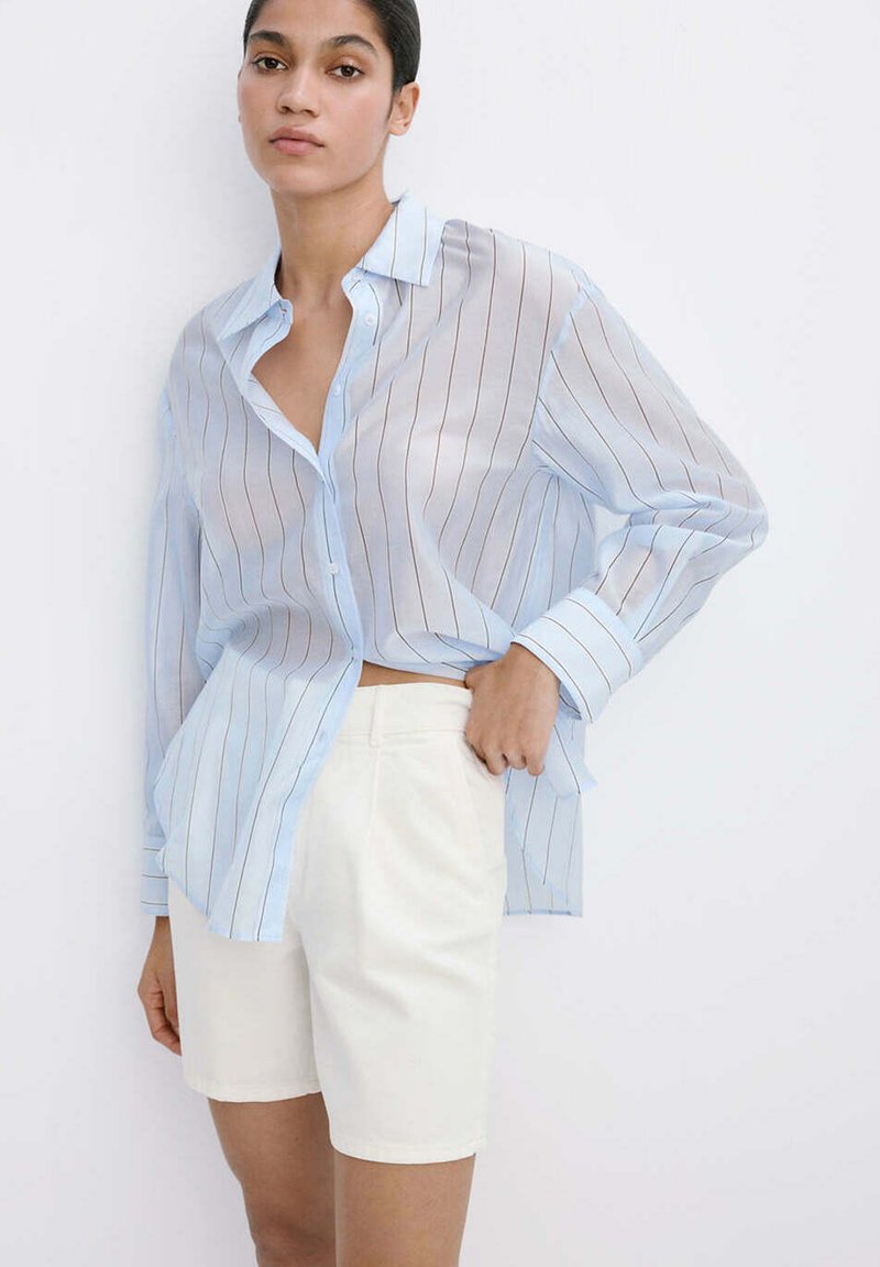 Femme portant une chemise bleu clair à manches longues à fines rayures, rentrée dans un short taille haute blanc cassé arrivant aux genoux, debout devant un fond blanc.
